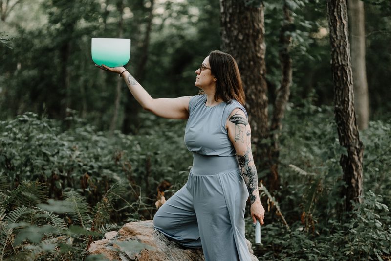 Mallory Bey kneels on a rock in the forest and holds a teal crystal singing bowl up to the light