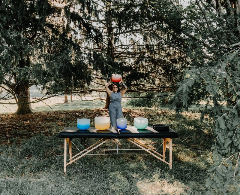 Mallory Bey stands behind her treatment table in a forest clearing holding a crystal singing bowl overhead, a row of bowls on the table