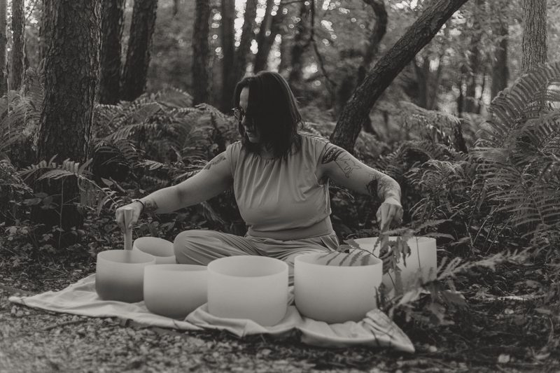 Black and white photograph of Mallory Bey kneeling beside a full set of crystal singing bowls laid out on a blanket among ferns