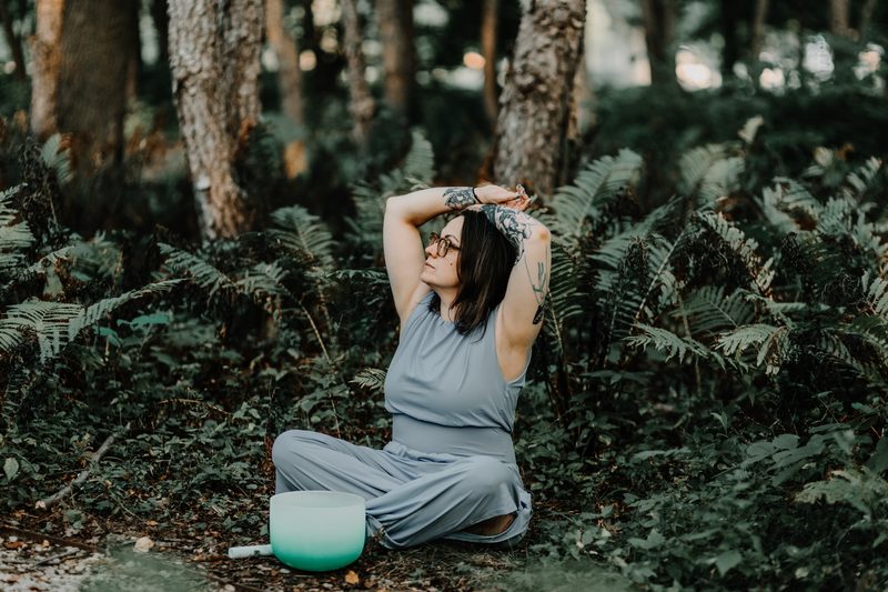 Mallory Bey sits among ferns in the forest with both arms stretched overhead, a teal crystal singing bowl beside her