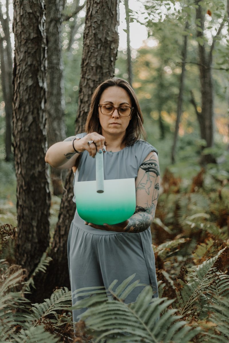 Mallory Bey stands among ferns in the forest playing a green crystal singing bowl