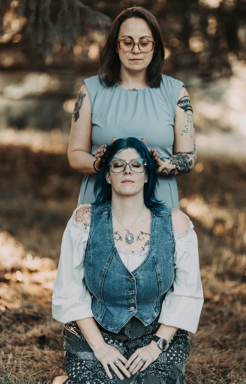 Mallory Bey stands behind a seated client in the woods with both hands resting on the client's shoulders