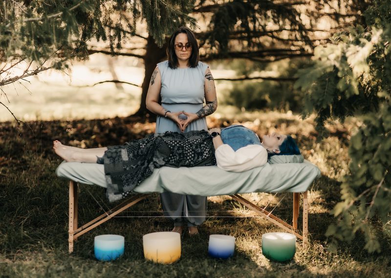 Mallory Bey sits at the head of the treatment table with her hands together while a client rests, four colored bowls in the grass