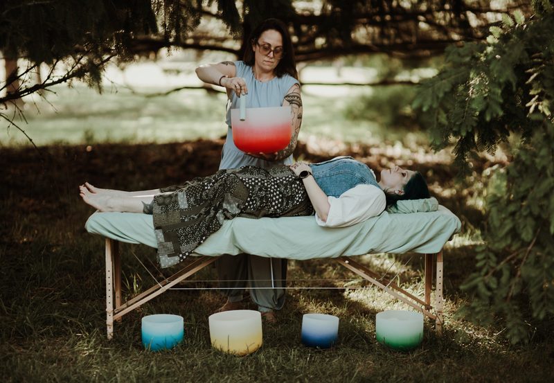 Mallory Bey plays a red crystal singing bowl while seated on the treatment table beside a reclining client, four bowls in the grass