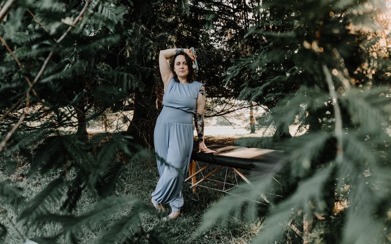 Mallory Bey stands among the pines beside her treatment table with one arm raised